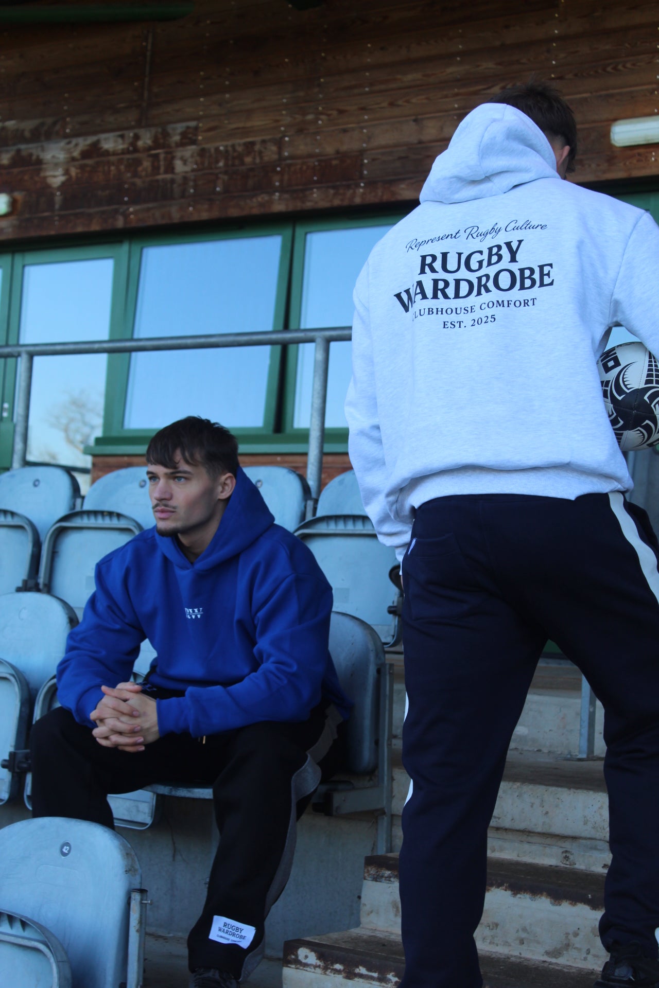 Two people sitting on bleachers with 'Rugby Wardrobe' branded represent rugby Hoodies.